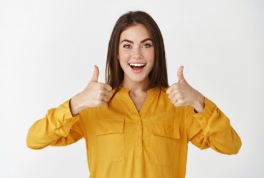 Impressed young woman showing thumbs up and smiling amazed, praising something cool, standing over white background.